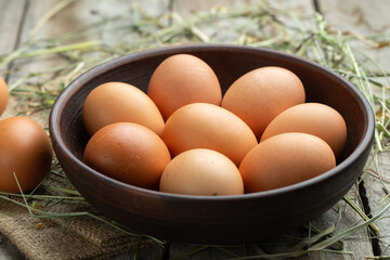 Chicken eggs in a clay bowl on a wooden background, hay scattered around. Healthy food concept.