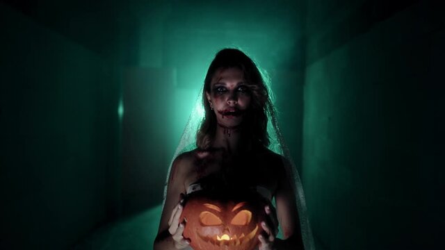 Zombie woman holding pumpkin and shouting close-up. Creepy dead bride with jack-o-lantern, wearing wedding dress. Bloody scary smile. Deceased fiancee returned to life. Halloween holiday.