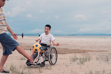 Happy disabled teenage boy on wheelchair playing a ball, Activity outdoors with father on the beach...
