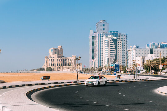 Toyota Taxi Car Moving In Street Of City Of Ajman. Building Of Bahi Ajman Palace Hotel In Sunny Day.Urban Architecture Of UAE Resort Town Of Ajman. View Of Cityscape