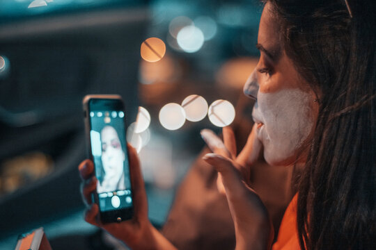 Adolescente joven pint&aacute;ndose por la noche mientras se observa en el tel&eacute;fono m&oacute;vil en un coche sentada con iluminaci&oacute;n de la ciudad
