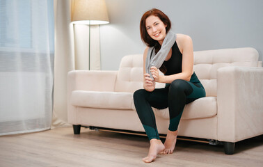 Female athlete is sitting on the couch resting after a fitness workout at home with a towel from sweat. Concept of weight loss.