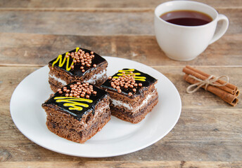 Pieces of cake, in the center of the frame, a kettle of tea in the distance on a dark background. Sweets for hot drinks. Three pieces of chocolate cake on a white plate.