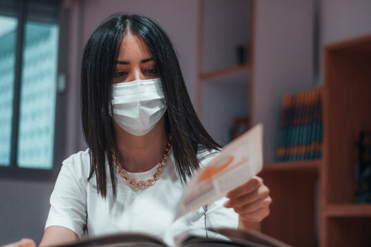 Joven adolescente con mascarilla pasando las paginas para leer su libro de biología en la sala de la biblioteca de su instituto