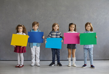 Team of 4 cute little kids showing colorful blank mock up sign boards or posters. Group of four happy children standing in grey studio and holding yellow, blue, pink and green mockup paper banners