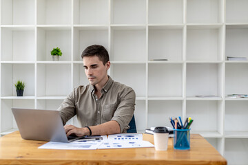 A businessman sits in his office with a laptop and stationery, on his desk there are financial documents he is checking, he is typing, texting, talking to his partner through a laptop messenger.