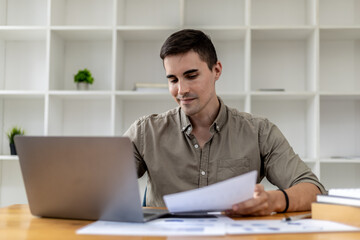 Fototapeta premium A young businessman sitting in the office working with a laptop, he is checking financial documents, he founded a startup, he is a young entrepreneur who has the skills to run a growing company.