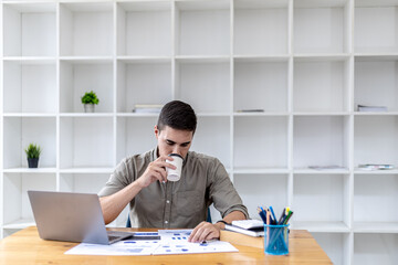 Fototapeta premium Young businessman sitting in the office with a laptop, taking a hot coffee break while checking documents, he founded a startup, he is a young entrepreneur who has the skills to run a growing company.
