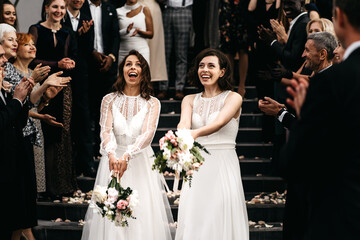 Candid shot of two female lesbian LGBT brides toss their bouquets over their shoulders to their...