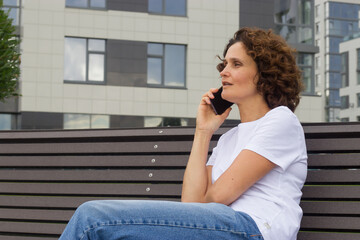 A 45-50-year-old woman is talking on a mobile phone while sitting on a bench