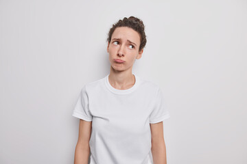 Depressed young sad European woman purses lips has gloomy face expression dressed in basic casual t shirt tilts head poses against grey background. Negative human emotions and feelings concept