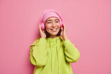 Pretty teenage girl listens music via headphones while strolling smiles positively wears hat casual hoodie poses against pink background. Female meloman listens songs downloaded from multimedia app