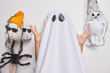 Autumn holiday. Faceless person pretends being spooky ghost covered with white sheet raises palms poses against scary decor indoor frightens you during night. Halloween party and celebration