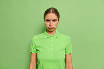 Unhappy frustrated young woman has sulking expression offended with someone frowns eyebrows dressed in casual t shirt isolated over vivid green background. Negative human emotions and feelings