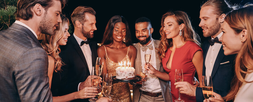 Group of people in formalwear holding cake with candles while celebrating birthday among friends