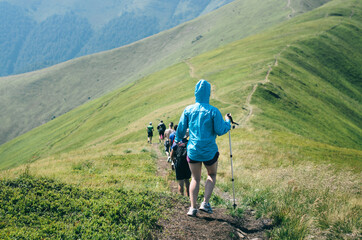 Back view of people go down the mountain ridge. The girl in a blue raincoat with a hiking stick goes behind the others.