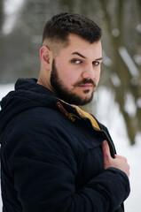 A young man with mustaches and raised eyebrow holds his hand over his jacket and walks in a pine forest. Portrait of a brutal bearded man. Winter forest tourism concept