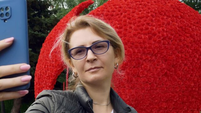 Stylish blonde woman in glasses makes a selfie on the background of a beautiful bouquet of red roses in the shape of a heart. A walk in the park with a smartphone and social networks.