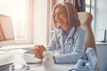 Beautiful mature female doctor in white lab coat smiling and looking away while sitting in her office