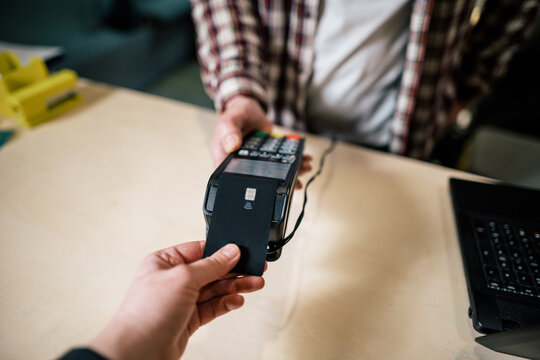 Close-up shopper hand gives credit card to pay through payment terminal, seller behind counter makes payment and gives goods in box to buyer. Pay use non-cash technology