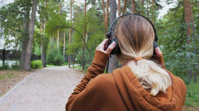 A young beautiful girl is running in a city park. She puts headphones on her head and listens to music while jogging. The camera shoots the blonde from the back. Active recreation and outdoor sports.