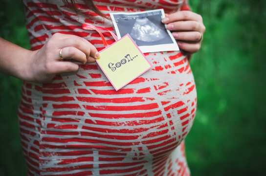 Pregnant woman holding a card soon and ultrasound scan or picture on the belly of her future daughter or son in the arms. Girl expecting newborn. Motherhood concept. Baby Shower.