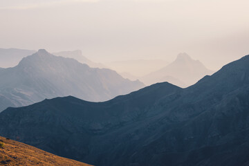 Scenic dawn mountain landscape with light fog in valley among mountains silhouettes under cloudy sky. Vivid sunset or sunrise scenery with low clouds in mountain valley in soft color.