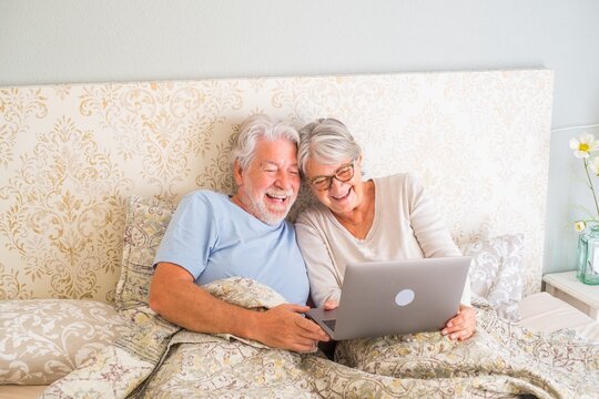 Old Senior Caucasian Couple Laughing And Using Laptop In The Morning At Bed In The Bedroom At Home. Elderly Couple Surfing And Using Social Media On Laptop At Home.