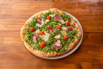 Arugula pizza with cherry tomatoes, parmesan cheese flakes, pieces of prosciutto and olive oil on wooden table