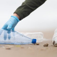 Plastic bottles on the sandy shore of the ocean A volunteer's hand in a glove collects bottles. Close-up. Poisoning with wildlife toxins. Ecological catastrophy. Garbage disposal.