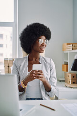 Beautiful young African woman using smart phone and smiling while sitting at her working place in...