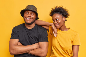 Obraz premium Indoor shot of two friendly woman and man with dark skin spend free time together being in good mood wear casual t shirts isolated over yellow wall. Afro American girl leans on shoulder of boyfriend