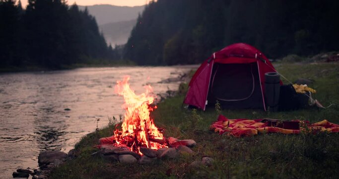 dancing flames of a campfire with tent, river and forest mountain on the background. Family travelling and camping concept