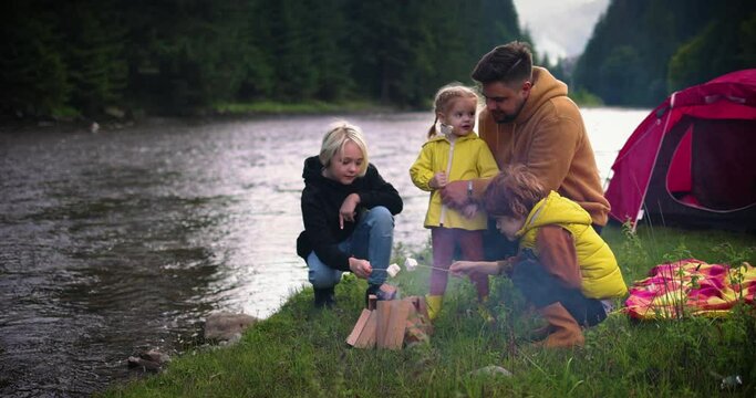 family with kids baking marshmallows on a campfire near the tent, with mountain forest and river at the background, active leisure