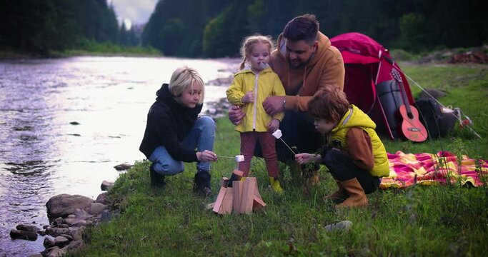 family with kids baking marshmallows on a campfire near the tent, with mountain forest and river at the background, active leisure