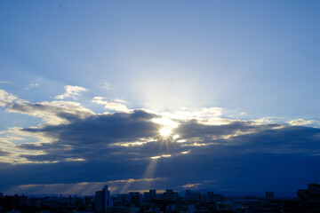 都市の夜明け。雲の隙間から太陽が顔を出し、天使の梯子が地上に降りる