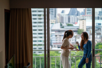 Millennial Asian woman friends standing on apartment balcony with drinking a cup of coffee and talking together. Confidence female friendship relax and enjoy weekend activity lifestyle at home. - obrazy, fototapety, plakaty