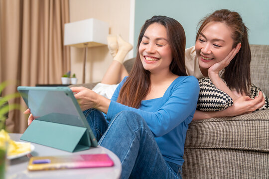 Asian Woman Friends Sitting In Living Room Using Digital Tablet For Watching Movie Or Social Media Together. Modern Female Friendship Enjoy Weekend Activity Lifestyle With Wireless Technology At Home.