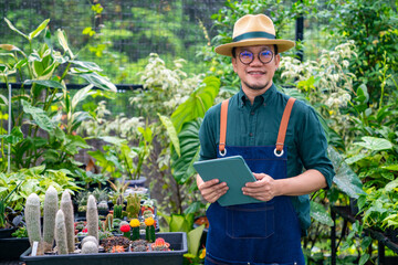 Asian man gardener caring plants in the garden. Male plant shop owner working on digital tablet for checking plants order in store. Small business entrepreneur working with technology concept.