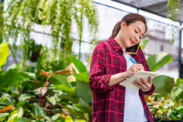 Asian woman gardener caring houseplants and flowers in greenhouse garden. Female plant shop owner taking order from customer on mobile phone. Small business entrepreneur and plant caring concept