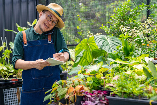 Asian Man Gardener Caring Houseplants And Flowers In Greenhouse Garden. Male Plant Shop Owner Taking Order From Customer On Mobile Phone. Small Business Entrepreneur And Plant Caring Concept