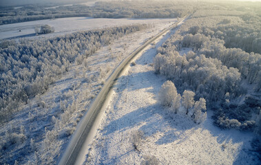 Aerial photo of winter road surrounded by birch forest. Drone shot of trees covered with hoarfrost and snow.