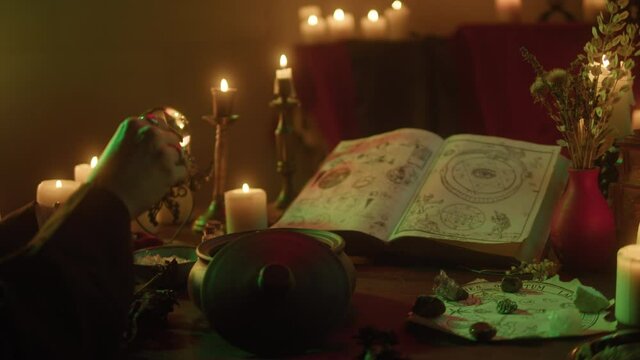 Witch cooking potion elixir close-up. Table with spellbook for making rite. Herbs, burning candles, crystal magic ball for sacrifice. Halloween theme, full moon, witchcraft. 