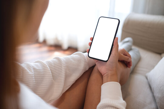 Mockup image of a woman holding mobile phone with blank desktop screen while sitting on a sofa at home