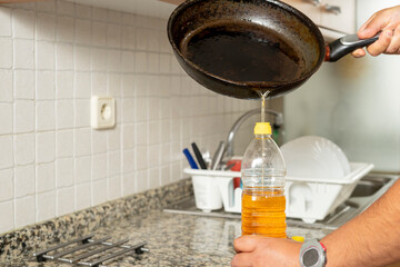 Man placing recycled edible oil from a frying pan into a plastic bottle in his home kitchen....