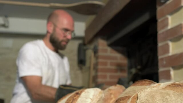 Close up side shot, baker&rsquo;s hand with gloves taking out loaf bread from the peel and placing it next to the traditional stone oven, on a rustic wooden table
