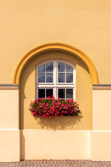 a white window placed in an arch-shaped recess against the background of a yellow wall. A flower pot with red geraniums on the window sill
