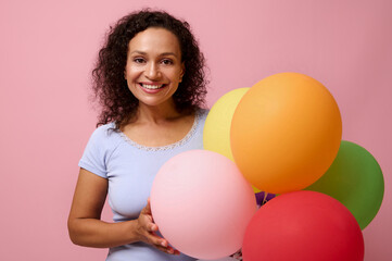 Fototapeta na wymiar Winsome attractive mixed race woman with a charming toothy smile, has happy reaction holding bright colorful helium balloons , celebrates birthday, looks at camera, isolated on pink colored background