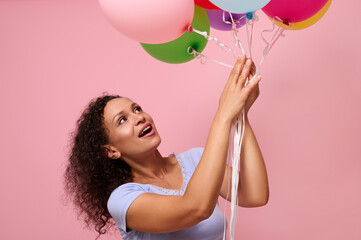 Studio shot for ad of a surprised woman reaching for colorful air balls smiling happily standing against pink background with copy space. The concept of celebration, holidays, anniversary and gifts.