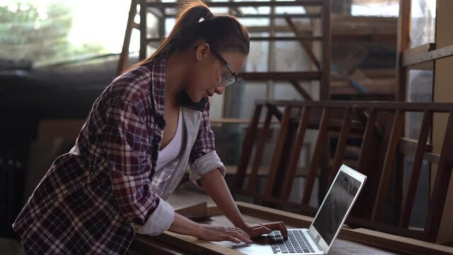 young asian business woman talking on phone making order on computer laptop in wood working factory . female carpenter working in her workshop
